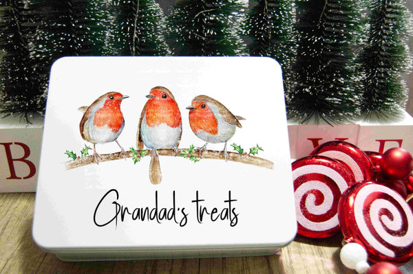 A white metal tin with a hand-drawn illustration of three robins on a branch with holly, titled 'Grandad's treats' on a wooden surface with Christmas decorations in the background.